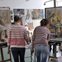 Dwie uczennice malują portret kobiety z modelu (widoczna kobieta w tle) w pracowni ZSP w Lublinie. Kliknięcie spowoduje wyświetlenie powiększonego zdjęcia.