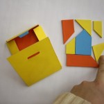 Układanka przypominająca puzzle o różnych kształtach w kolorach: żółty, czerwony, niebieski. Obok pudełko na układankę w tych samych barwach. Ktoś palcem wskazującym dopasowuje kształty układanki. Kliknięcie w miniaturkę obrazka spowoduje wyświetlenie powiększonego zdjęcia.