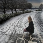 Młoda blondynka z długimi włosami ubrana w ciemny płaszczyk w pozyccji kucącej na drodze rowerowej. Jest zima, śnieg. w tle drzewa i żywopłot. Kliknięcie w miniaturkę obrazka spowoduje wyświetlenie powiększonego zdjęcia.