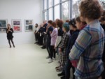 Marzena Łukaszuk przemawia na tle prac w galerii. Obok stoją ludzie, w większości młodzież. Kliknięcie w miniaturkę obrazka spowoduje wyświetlenie powiększonego zdjęcia.