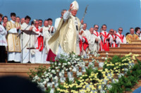 Scena dzieje się na dworze. Papież Jan Paweł II pozdrawia ludzi uniesioną dłonią. Za nim stoi dużo duchownych. Niżej na schodach kwiaty białe i żółte. Kliknięcie w miniaturkę obrazka spowoduje wyświetlenie powiększonego zdjęcia.