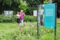 Dwie kobiety oglądają wystawę na terenach zieleni wokół szkoły. Jedna z nich fotografuje. Kliknięcie w  miniaturkę obrazka spowoduje wyświetlenie powiększonego zdjęcia.