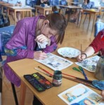 Uczennica rysuje i maluje sceny komiksu w auli szkolnej. Kliknięcie w miniaturkę obrazka spowoduje wyświetlenie powiększonego zdjęcia.