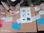 Uczniowie siedzą przy stołach i piszą na karteczkach. Obok nich leżą inne materiały z papieru, wydruki z tekstem, itp. Kliknięcie w miniaturkę obrazka spowoduje wyświetlenie powiększonego zdjęcia.