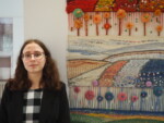 Uczennica stoi przy swojej pracy dyplomowej z tkaniny artystycznej zawieszonej na ścianie korytarza szkolnego. Kliknięcie w miniaturkę obrazka spowoduje wyświetlenie powiększonego zdjęcia.