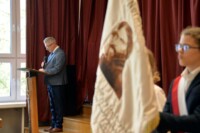 Dyr Krzysztof Dąbek przemawia stojąc przy mównicy w auli szkolnej. Uczeń trzyma sztandar szkoły. Kliknięcie w miniaturkę obrazka spowoduje wyświetlenie powiększonego zdjęcia.