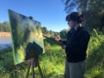 Ucznennica maluje obraz na płótnie umieszczonym na sztaludze w plenerze wśród drzew nad rzeką. Kliknięcie w miniaturkę obrazka spowoduje wyświetlenie powiększonego zdjęcia.