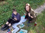 Nauczycielka Anna Chmielnik ochyla się nad chłopcem, który siedzi na materiale na ziemi. Obok niego przybory malarskie. Kliknięcie w miniaturkę obrazka spowoduje wyświetlenie powiększonego zdjęcia.