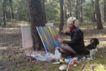 Dziewczyna maluje krajobraz w lesie. Obok niej siedzi pies. Kliknięcie w miniaturkę obrazka spowoduje wyświetlenie powiększonego zdjęcia.