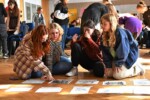 Uczniowie siedzą na podłodze i układają kartki z grafikami na podłodze w auli szkolnej. Kliknięcie w miniaturkę obrazka spowoduje wyświetlenie powiększonego zdjęcia.