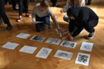 Uczniowie siedzą układają kartki z grafikami na podłodze w auli szkolnej. Kliknięcie w miniaturkę obrazka spowoduje wyświetlenie powiększonego zdjęcia.