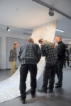 Trzej nauczyciele ubrani w koszule w kratkę i dyr Krzysztof Dąbek oglądają dużą, jasną pracę zwisającą i rozłożoną, jak dywan. Kliknięcie w miniaturkę obrazka spowoduje wyświetlenie powiększonego zdjęcia.