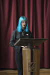 Uczennica z niebieskim włosami i z wisiorkiem (duży krzyż) na szui stoi przy mównicy i przemawia. Kliknięcie w miniaturkę obrazka spowoduje wyświetlenie powiększonego zdjęcia.