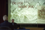 Adrian Lesiakowski siedzi przy stole i prowadzi wykład. W tle slajd na ekranie multimedialnym przedstawijący postacie historyczne i napis Długi wiek XIX. Kliknięcie w miniaturkę obrazka spowoduje wyświetlenie powiększonego zdjęcia.