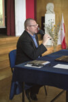 Adrian Lesiakowski siedzi przy stole przykrytym granatowym suknem i prowadzi wykład. W tle popiersie C. K. Norwida, flaga Polski i wazon z suchymi kwiatami. Kliknięcie w miniaturkę obrazka spowoduje wyświetlenie powiększonego zdjęcia.